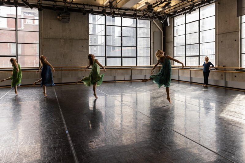 Four dancers in floaty green tunics crook one leg behind them and lean back to look back at the ceiling. Lori Belilove leans against a ballet barre as she watches them.